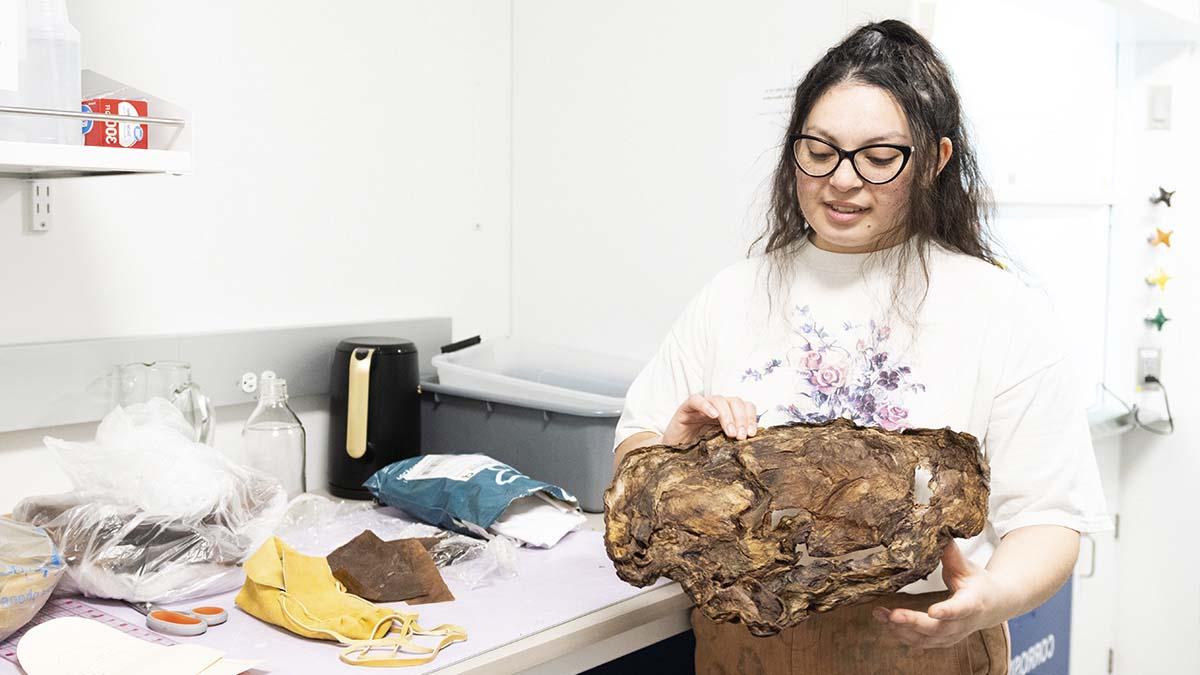 A woman holds a large, dried SCOBY in a lab next to a table with patterns, scissors and a leather moccasin.
